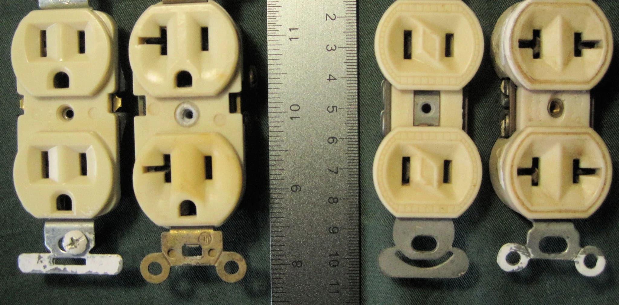 A 15-amp duplex receptacle beside a 20-amp duplex receptacle whose neutral slots have a sideways T-shaped notch, next to a metal ruler and two older two-slot receptacles on dark green cloth.