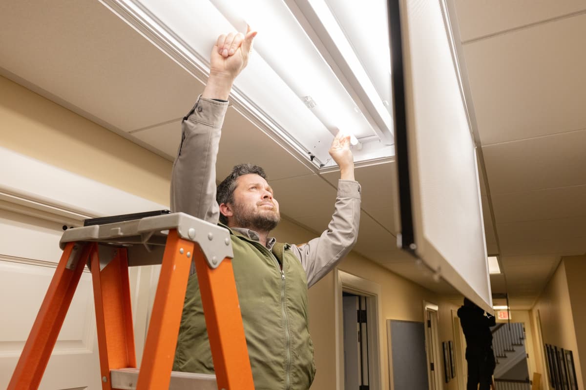 An electrician standing on an orange stepladder reaches overhead to install new LED lamps in a hallway ceiling light fixture.