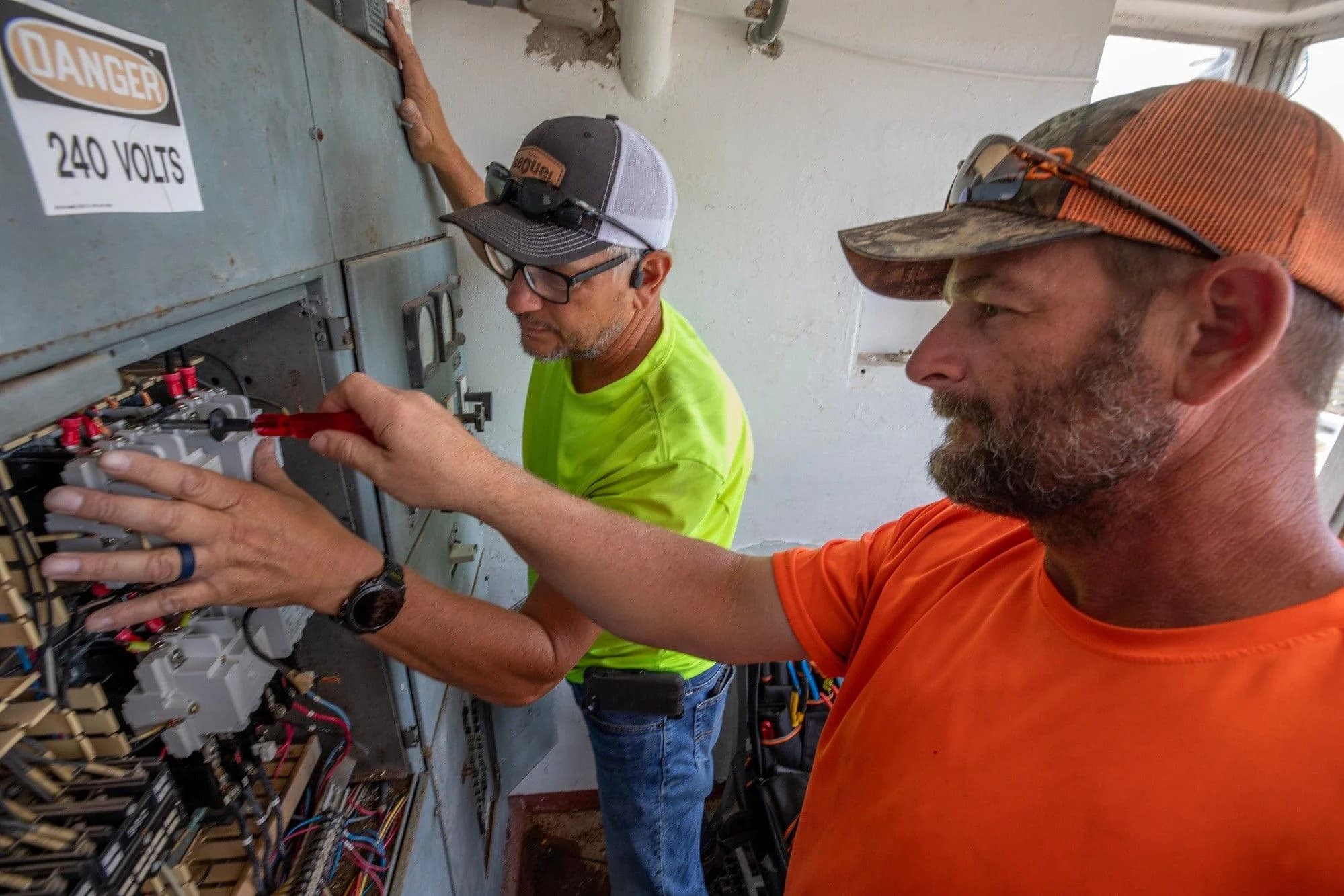 Two electricians in civilian work clothes stand at an open industrial control cabinet marked DANGER 240 VOLTS, one reaching in toward the terminal blocks with a screwdriver while the other steadies the panel.