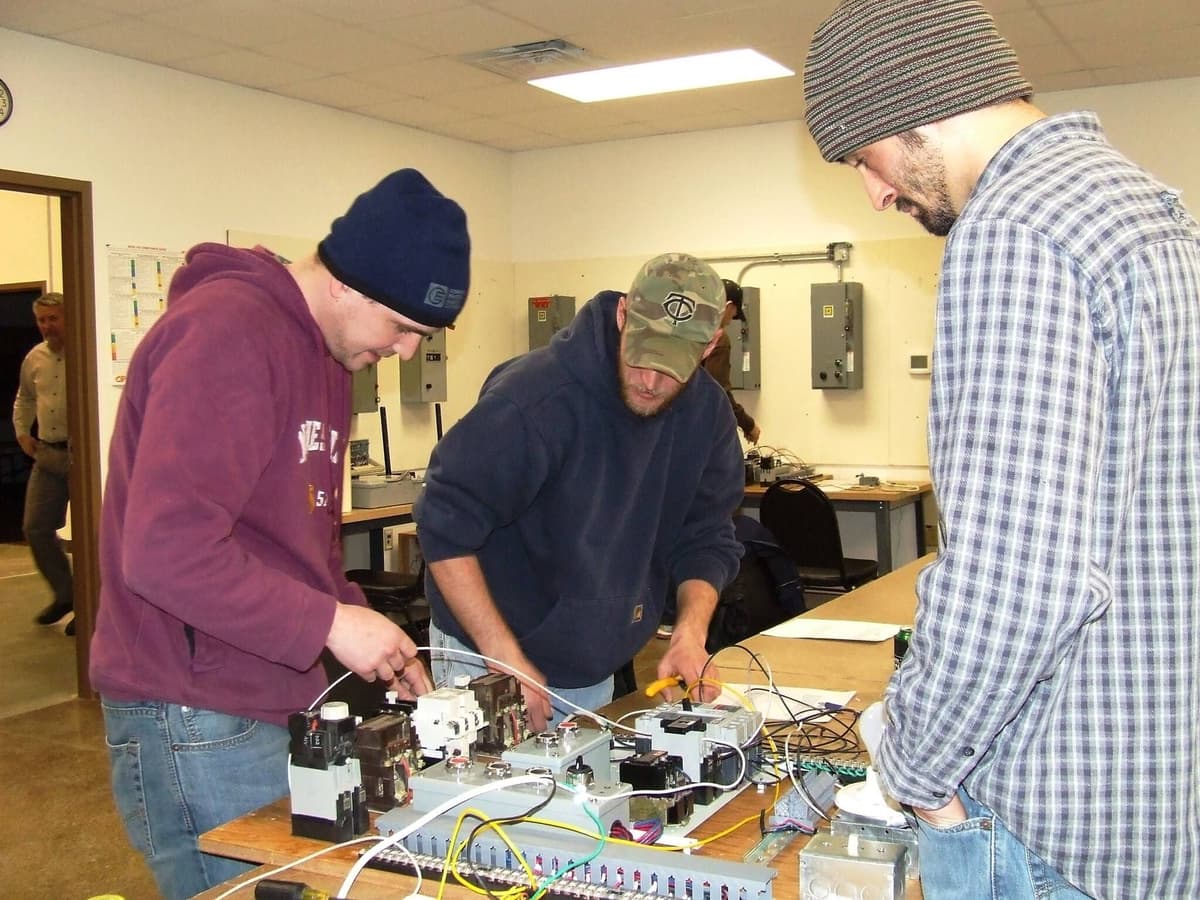 Three adult electrical apprentices wire a motor-control training board together at a workbench in a training-center classroom, with panelboards mounted on the wall behind them
