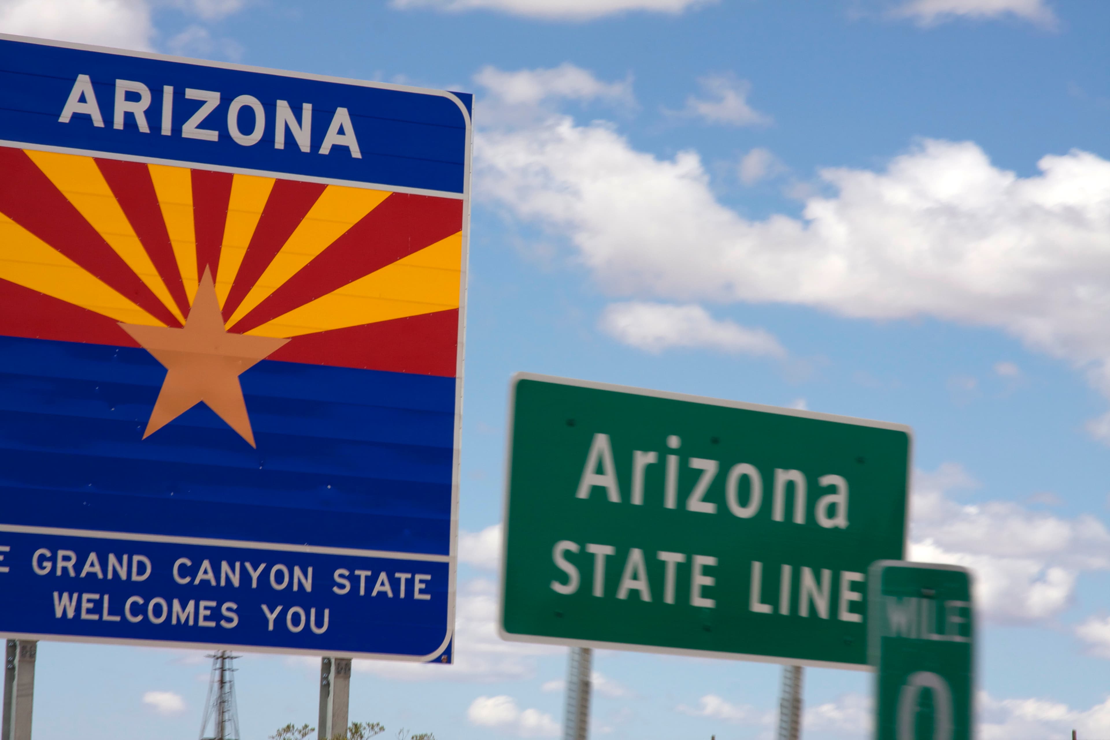 The blue Arizona welcome sign with the state flag's star and rays stands beside a green "Arizona State Line" highway sign and a Mile 0 marker on Interstate 10 under a blue sky.