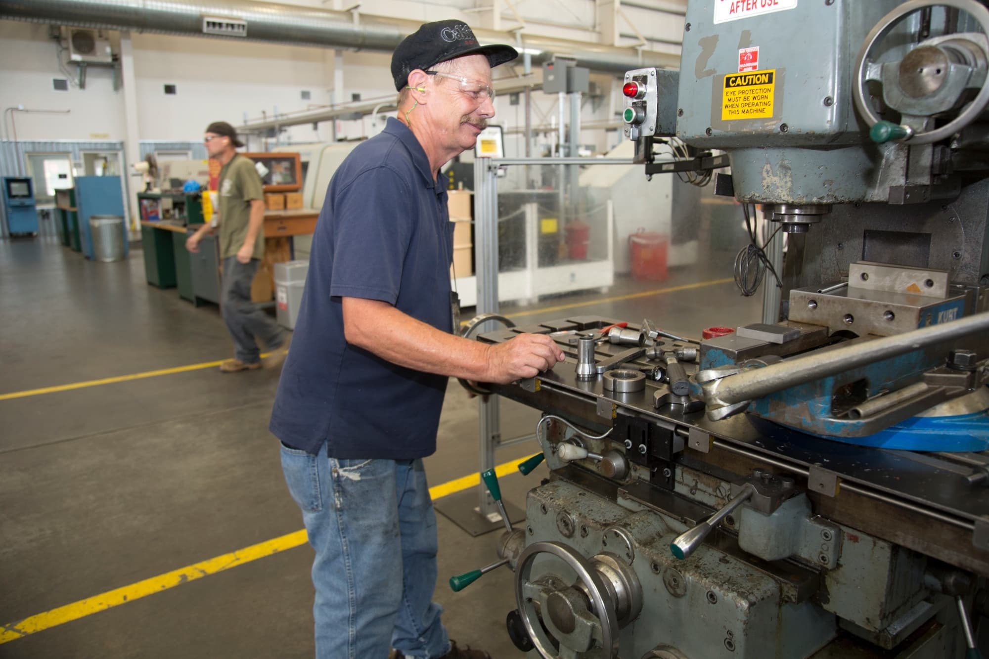 A veteran machinist in a ball cap, safety glasses and earplugs works at a vertical milling machine on a brightly lit American shop floor, with another worker and rows of machines behind him.