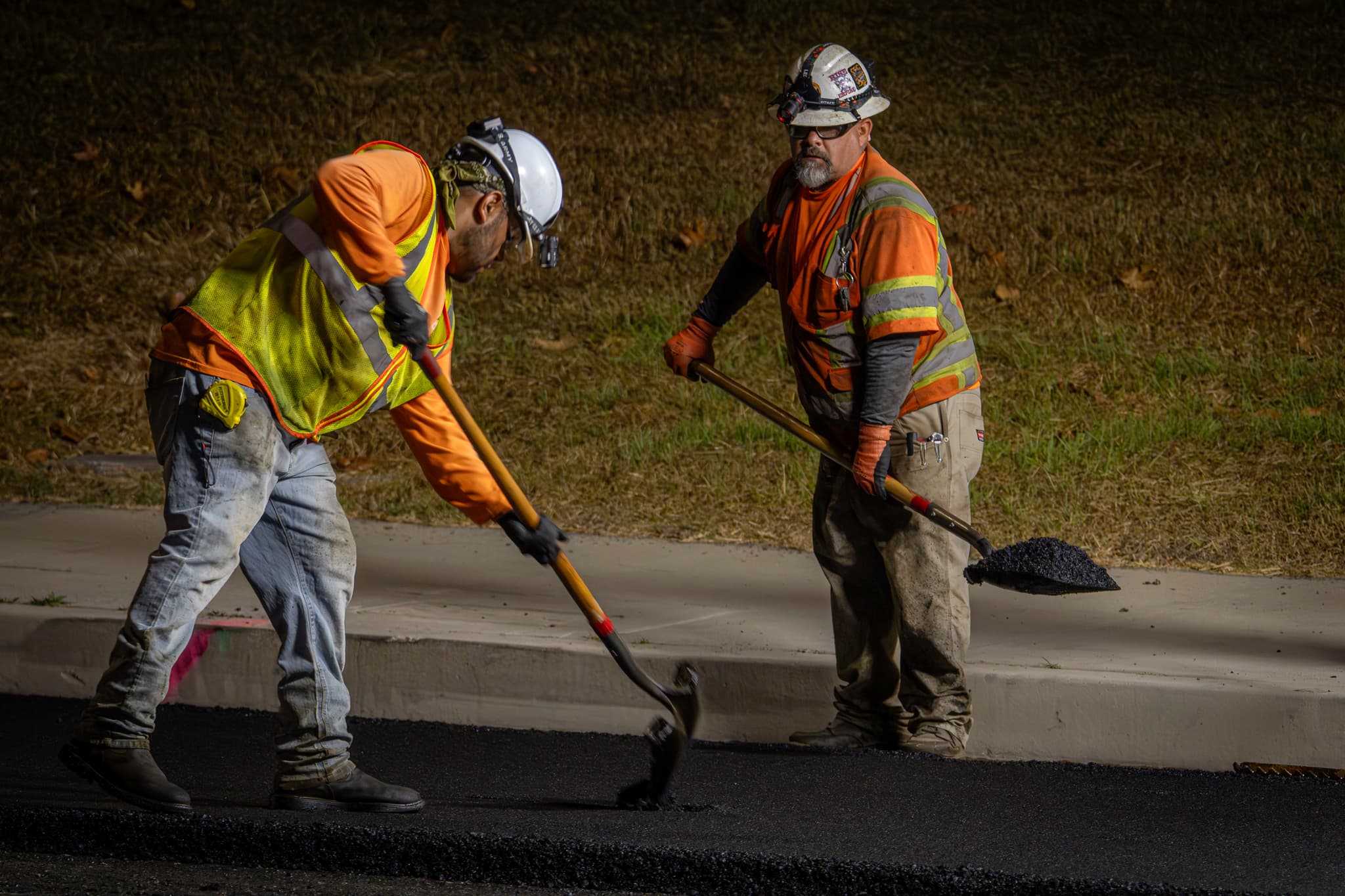 Two road workers in hi-vis shovel hot asphalt under work lights on a night paving shift.