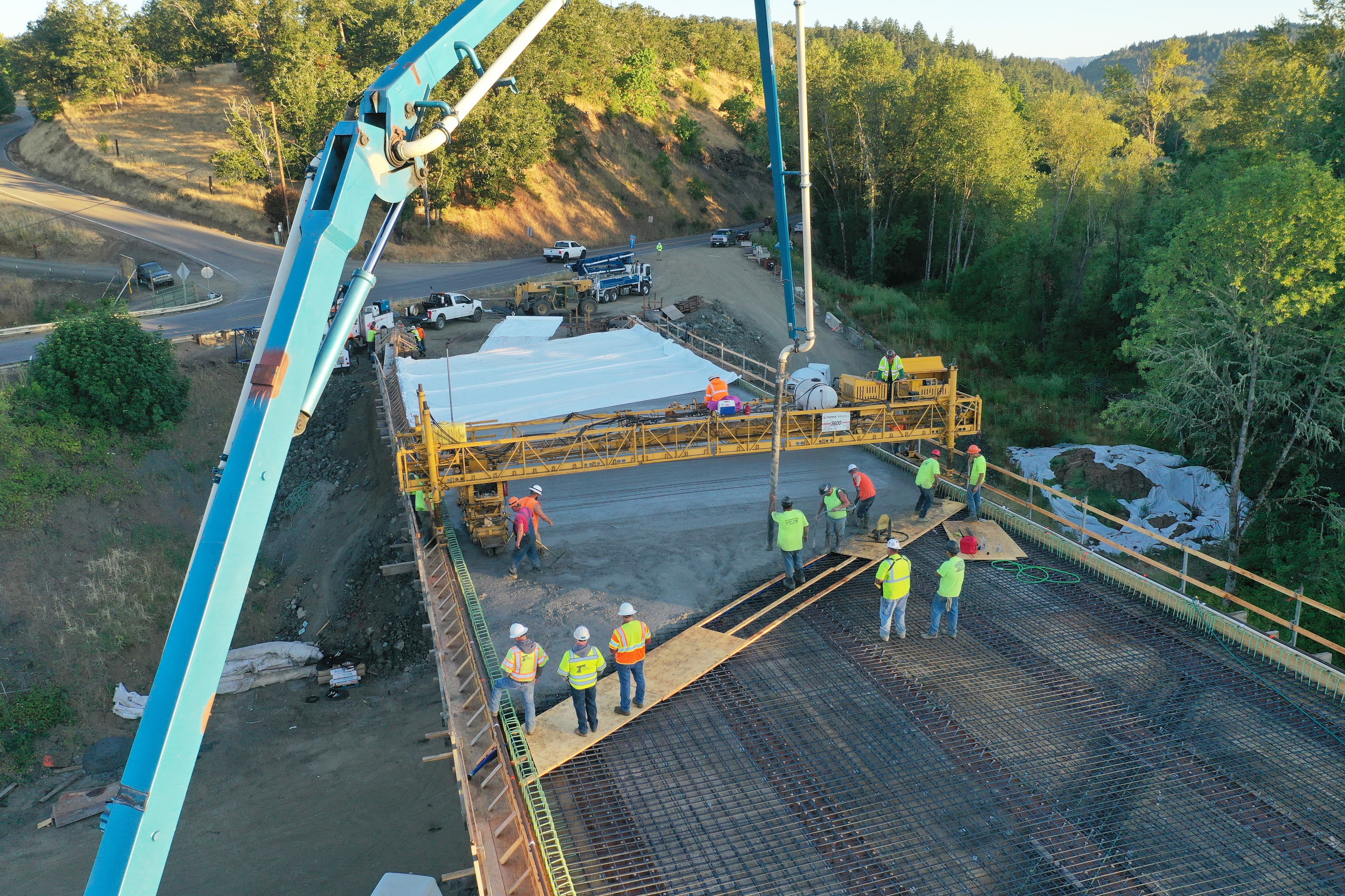 Aerial view of a bridge deck concrete pour with about fourteen hi-vis workers spread across the rebar and screed.