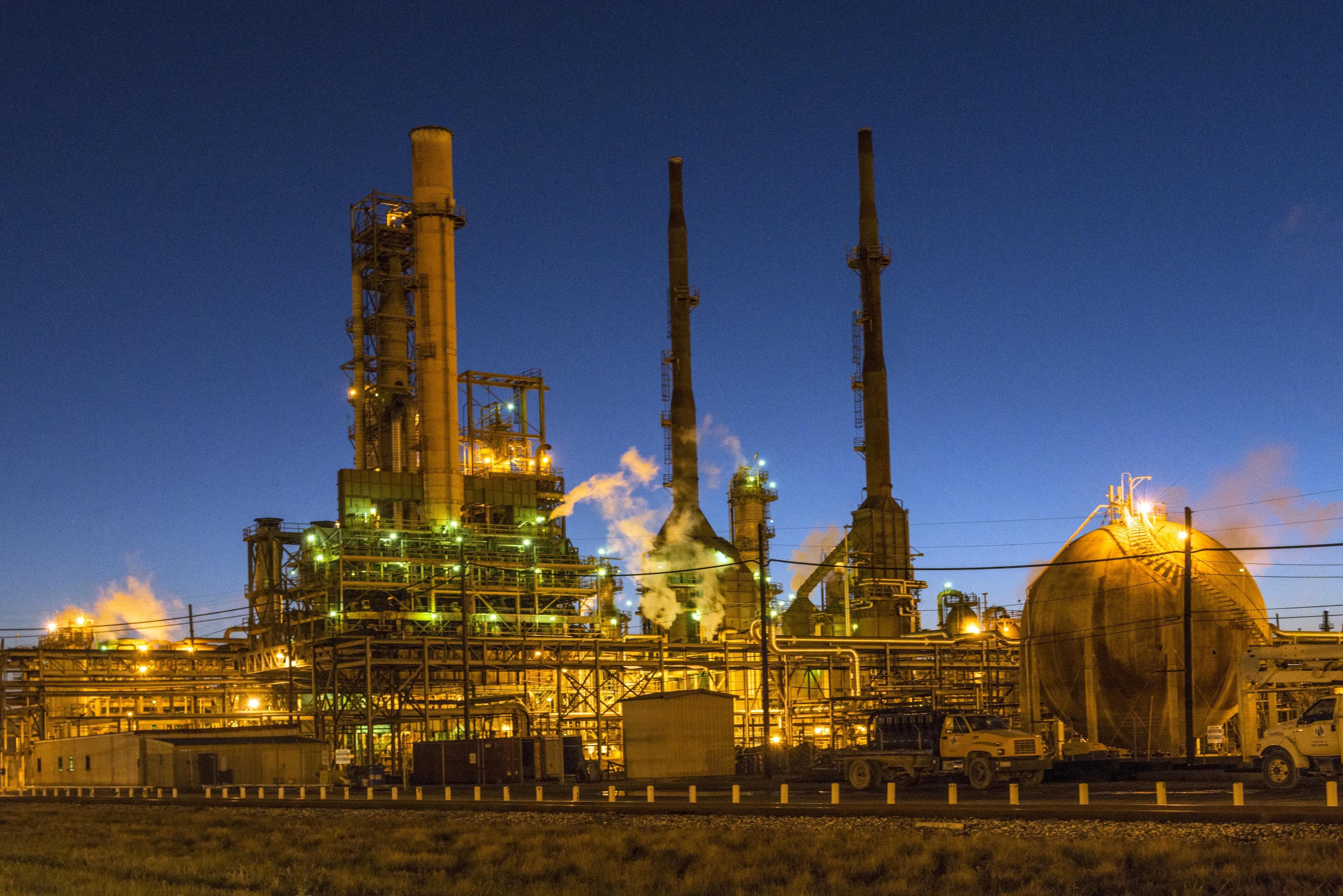 A Texas oil refinery lit up at dusk, its distillation columns, stacks and storage sphere glowing orange against a deep blue sky, with a pickup truck parked inside the unit fence.