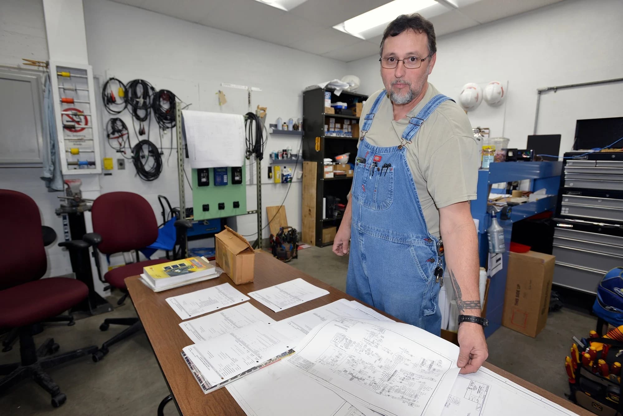 A veteran power plant electrician in denim bib overalls stands at his shop workbench with electrical wiring schematics spread out in front of him.