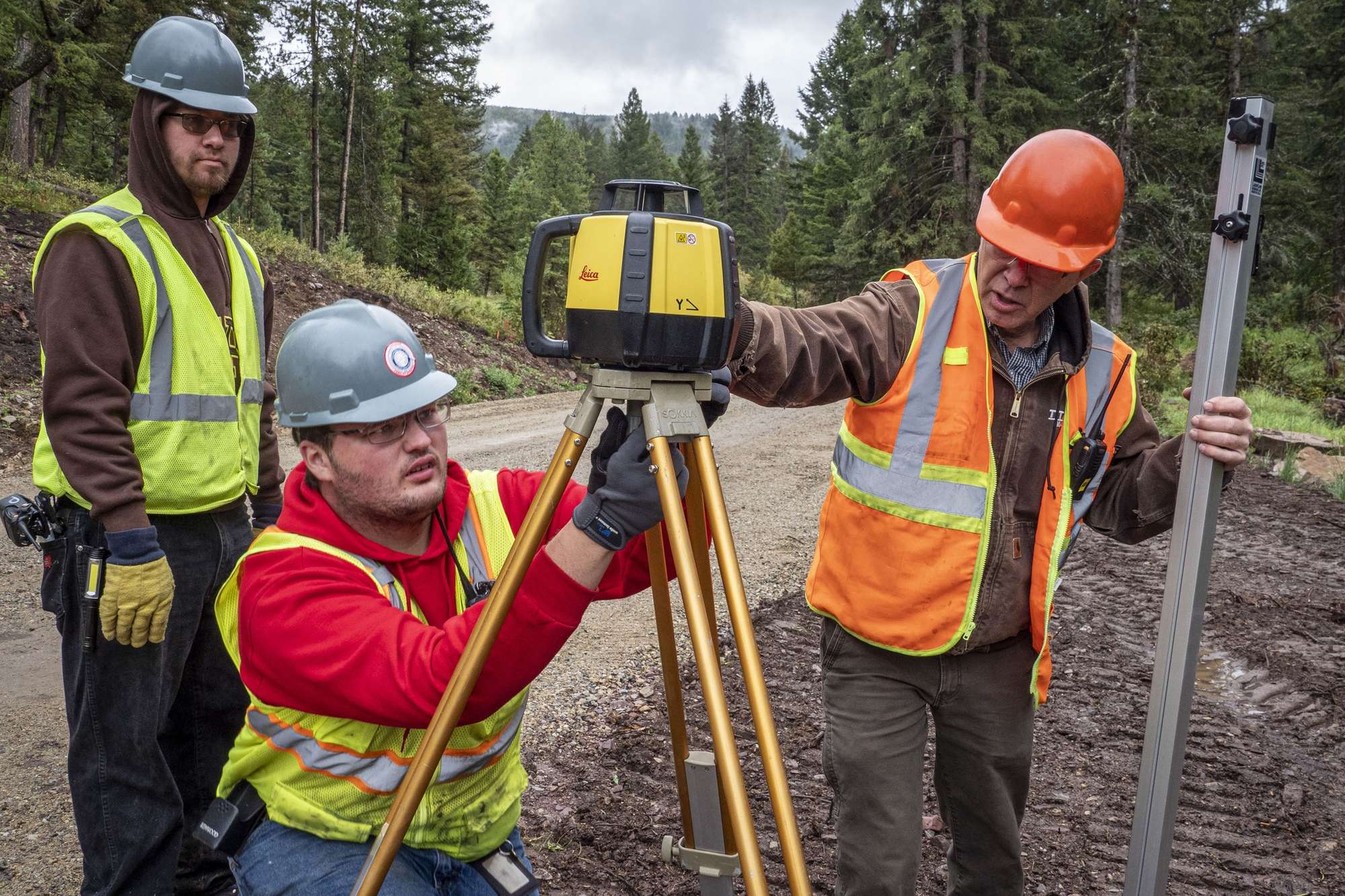 Two Job Corps students in hard hats and high-visibility vests set up a yellow rotating laser on a survey tripod while an instructor in an orange hard hat steadies a grade rod on a gravel forest road.