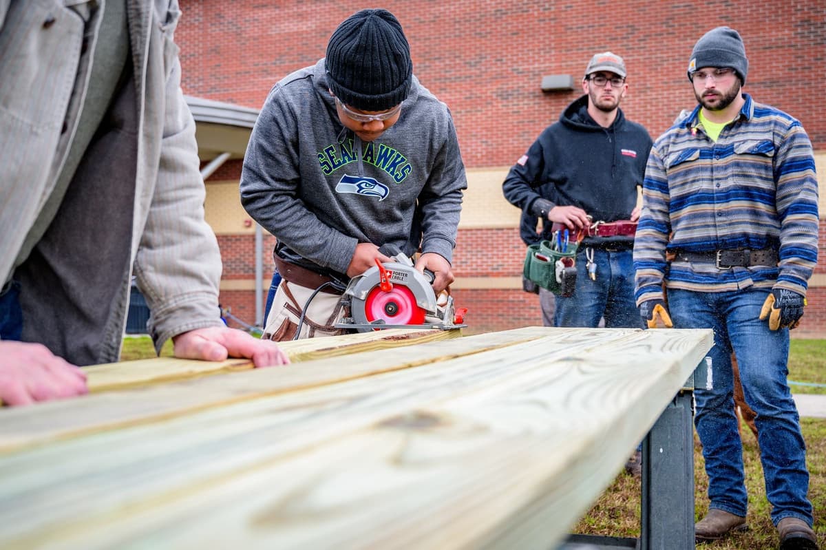 A trainee in safety glasses and a tool belt cuts a treated board with a circular saw while two classmates in beanies and tool belts watch at an outdoor construction workforce training session.