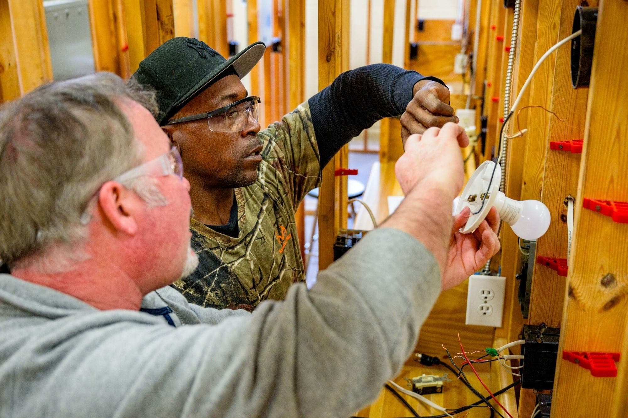 An instructor in safety glasses leans in beside a trainee who is wiring a lampholder on a framed training wall in a community college electrical lab.