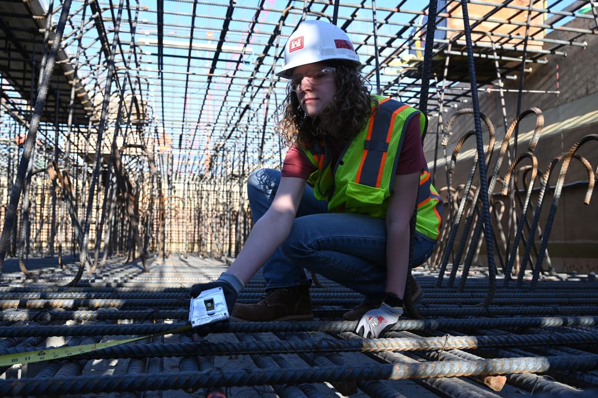 A construction worker in a hard hat, safety glasses and hi-vis vest kneels with a tape measure on a dense two-layer rebar mat, with a tunnel of vertical bars and hooked dowels receding behind