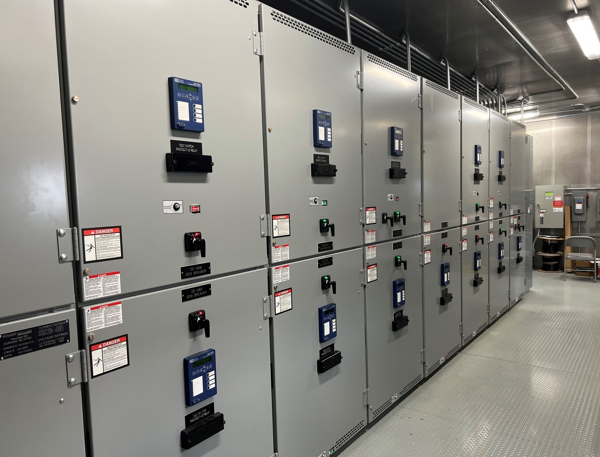 A long receding row of gray medium-voltage electrical switchgear cabinets in a clean facility interior, fitted with digital protective relays, rotary breaker handles, and red danger placards.
