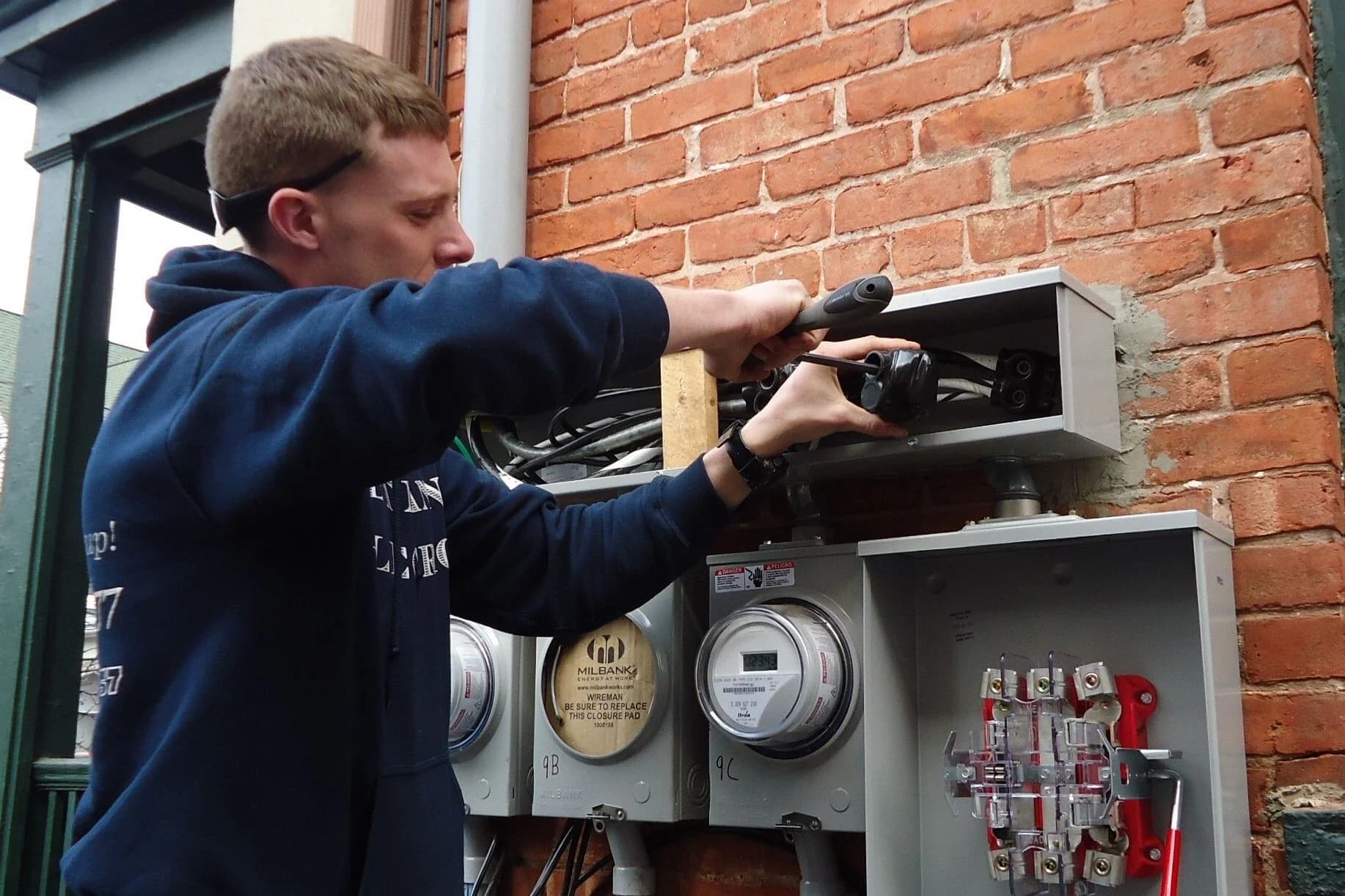A young electrician in a navy hooded sweatshirt reaches into an open wiring trough above a bank of residential electric meters on a brick wall, working a heavy insulated cable connector.