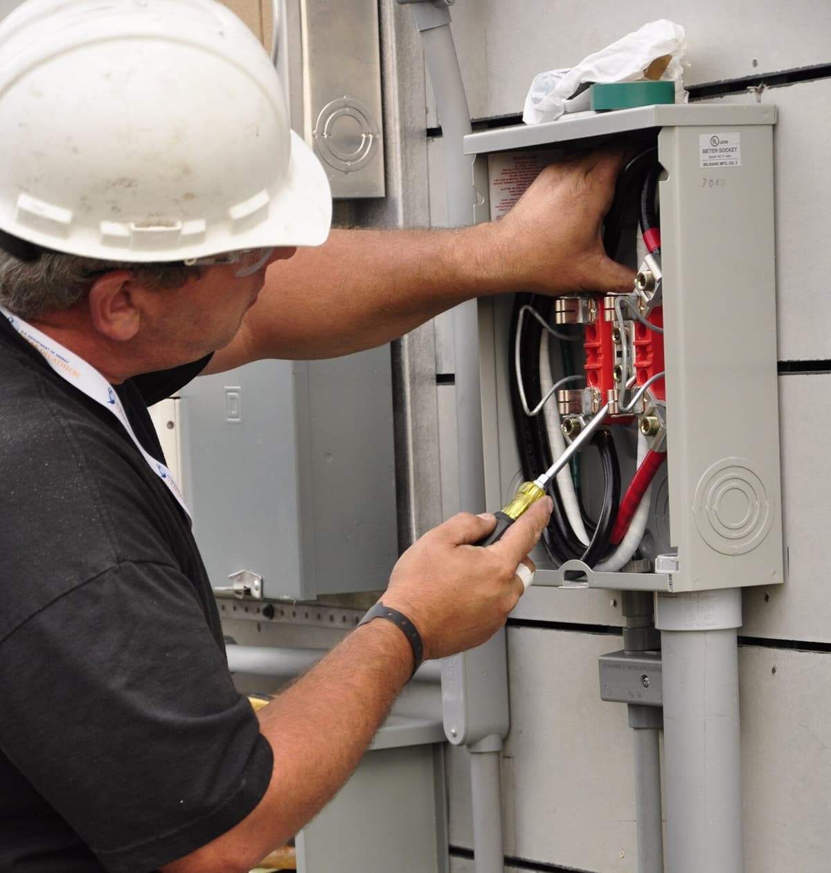 An electrician in a white hard hat and safety glasses reaches into an open meter socket on the side of a house, driving a lug screw with a screwdriver while steadying the service conductors with his other hand.