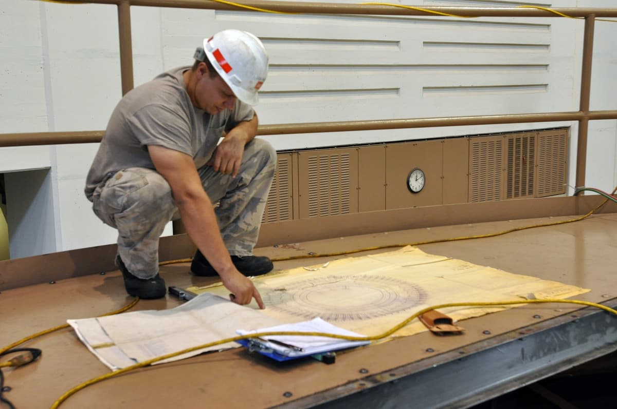 An electrician in a white hard hat and gray T-shirt crouches on a generator deck and points to a yellowed paper drawing with a large circular diagram, spread on the floor beside a clipboard.