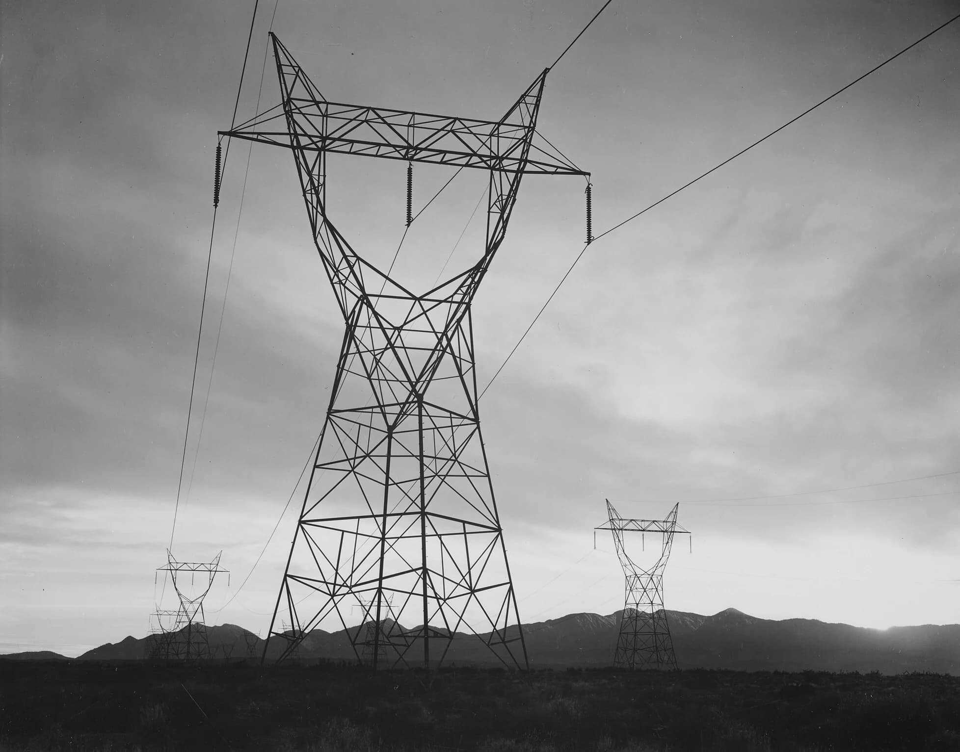 Black-and-white 1941 photograph of a tall steel lattice transmission tower with long insulator strings in the Mojave Desert, more towers receding toward mountains under a cloudy sky