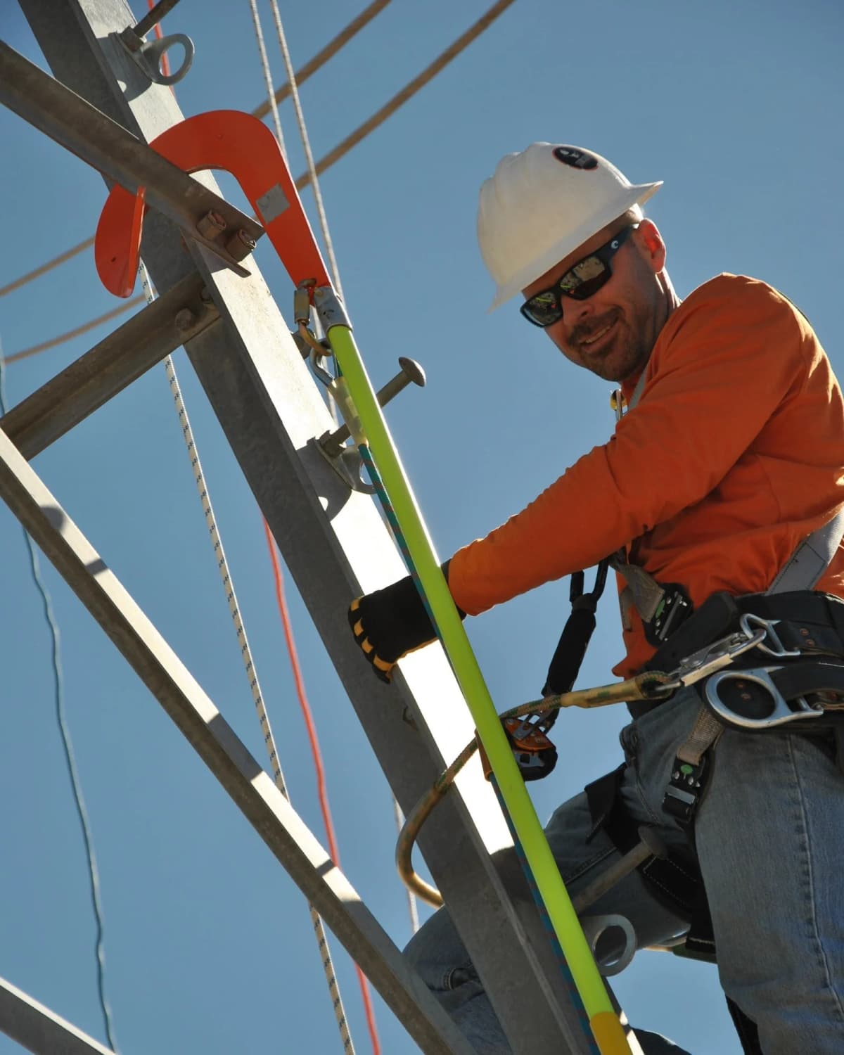 A smiling lineman in a white hard hat and orange shirt climbs a steel lattice transmission tower using a buck hook and fall-arrest line