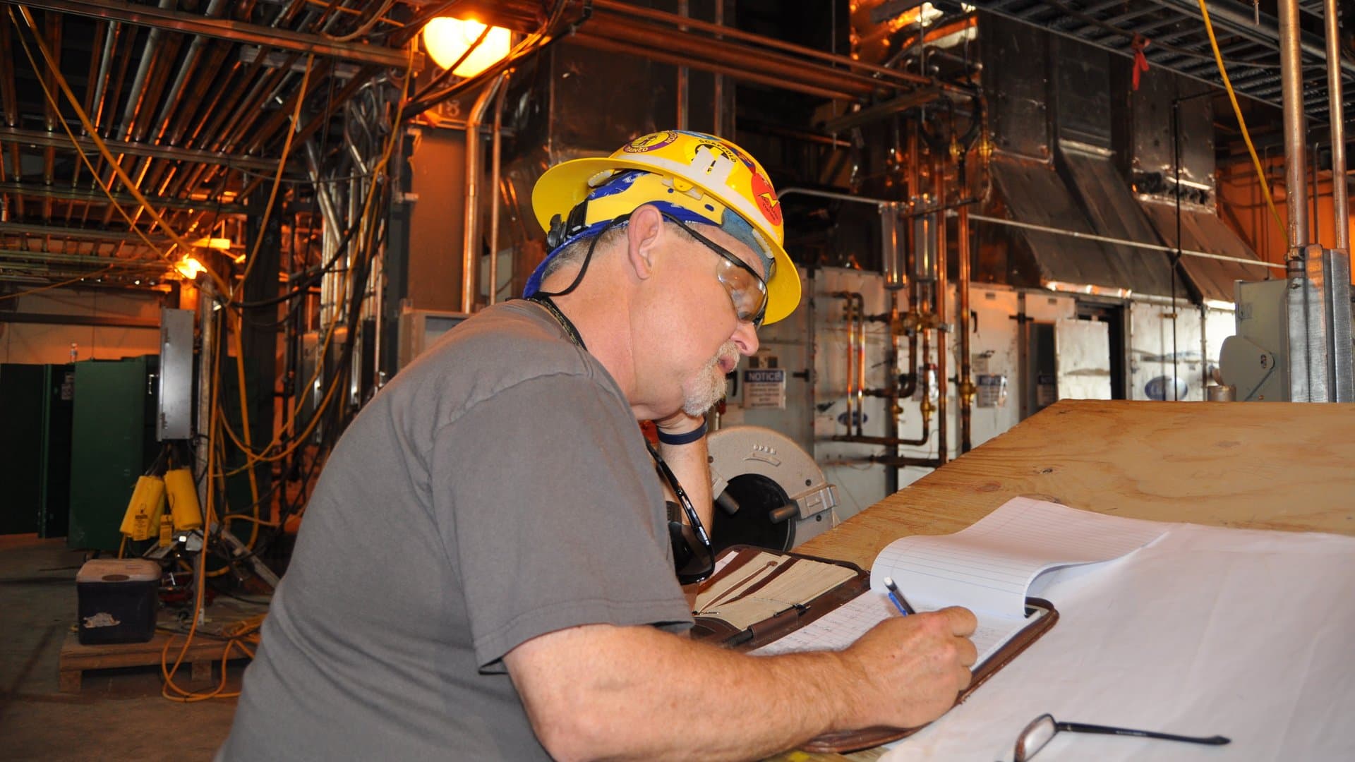 An electrician in a yellow hard hat and safety glasses writes notes in a padfolio at a plywood work table in a mechanical room.