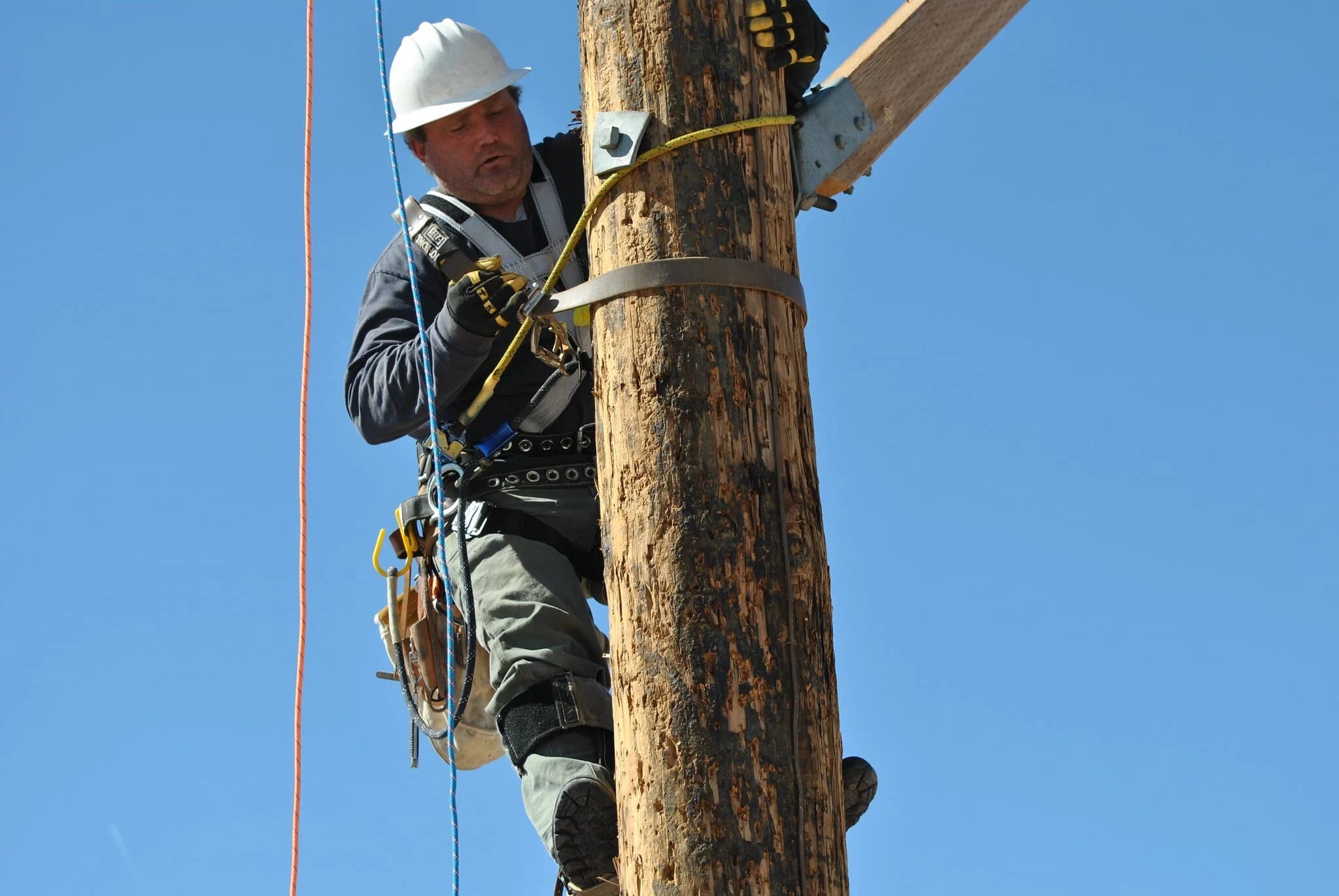 A lineworker in a white hard hat and full-body harness grips a wooden utility pole near a crossarm, secured by a pole strap and body belt, against a clear blue sky.