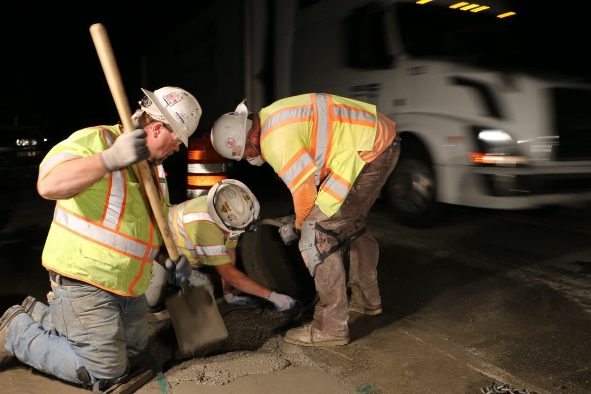 Three road workers in high-visibility vests and hard hats kneel over a fresh concrete deck patch at night, one holding a tamping bar and one a shovel, as a semi truck passes in the open lane beside them.