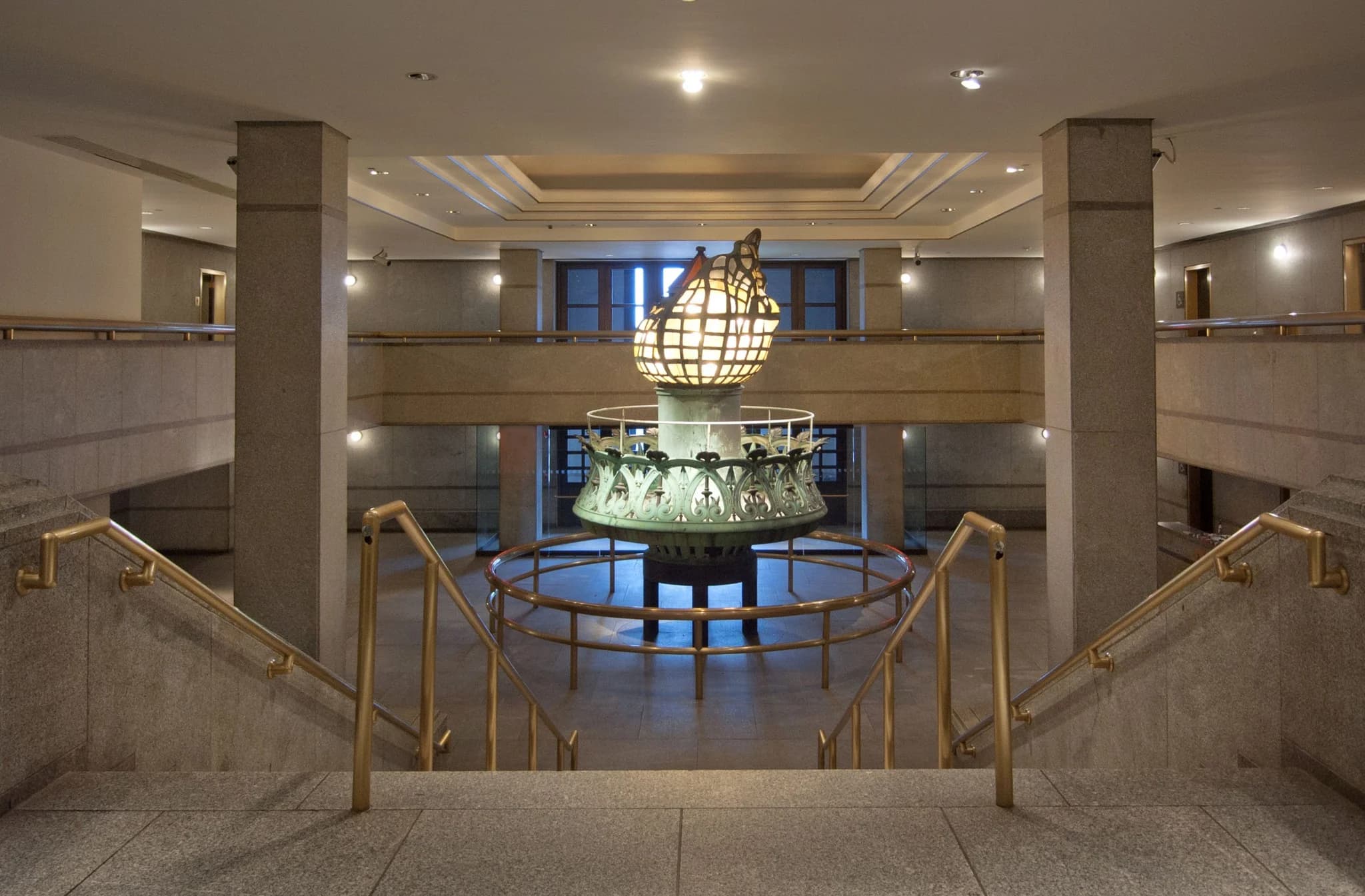 The original Statue of Liberty torch on exhibit in the pedestal lobby, its glass-paned flame glowing amber above the green copper balcony, with brass stair railings in the foreground