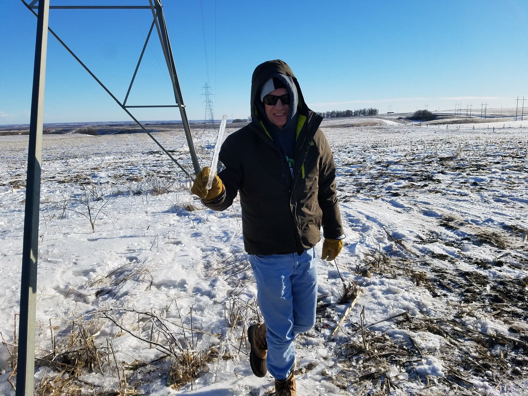 A smiling lineworker in a hooded winter parka, beanie, sunglasses and leather work gloves walks across an icy, snow-covered field beside a steel transmission tower, holding up a long piece of clear ice.