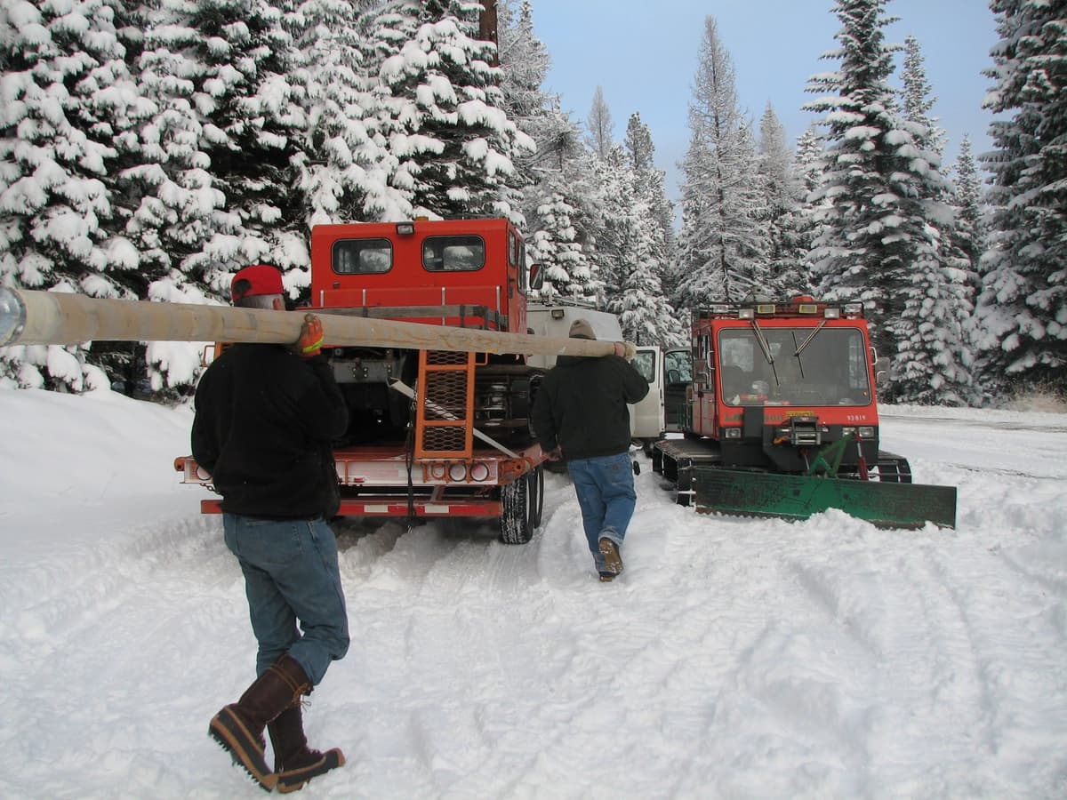 Two technicians in dark winter jackets, jeans and tall insulated boots carry a long antenna on their shoulders toward an orange snowcat on a trailer, with a second snowcat and snow-laden fir trees beside them.